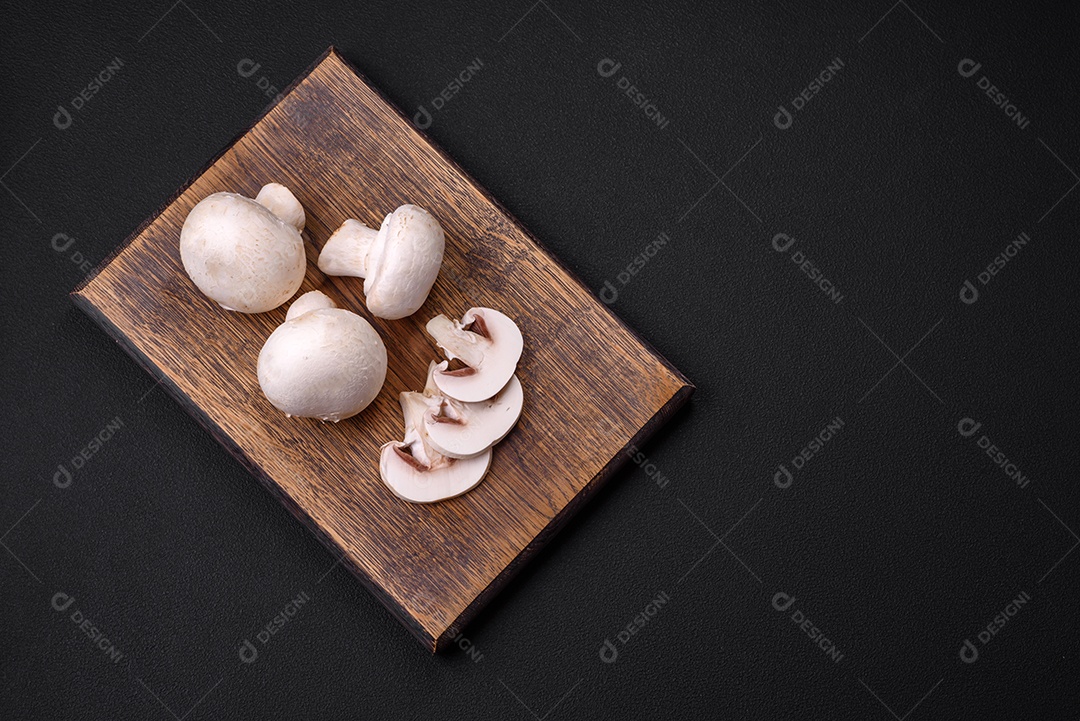 Cogumelos champignon crus frescos sobre uma tábua de madeira com especiarias e ervas sobre um fundo escuro de concreto