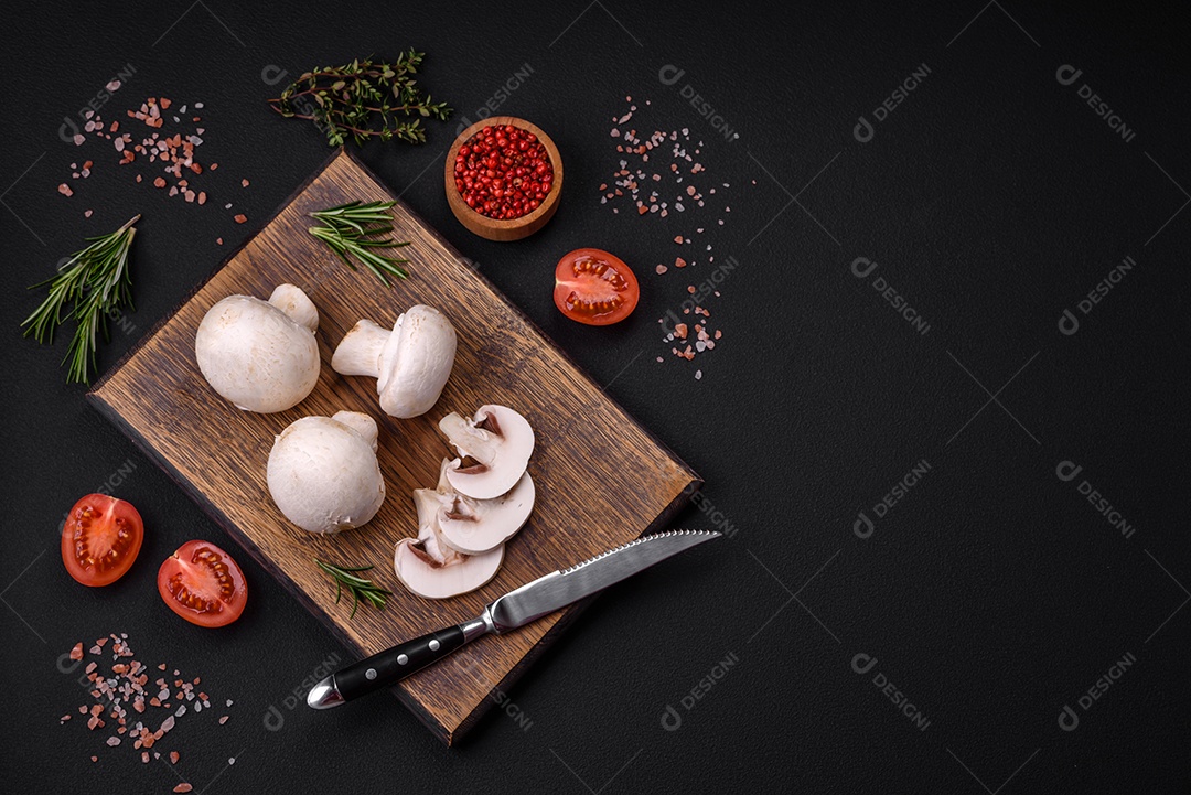 Cogumelos champignon crus frescos sobre uma tábua de madeira com especiarias e ervas sobre um fundo escuro de concreto