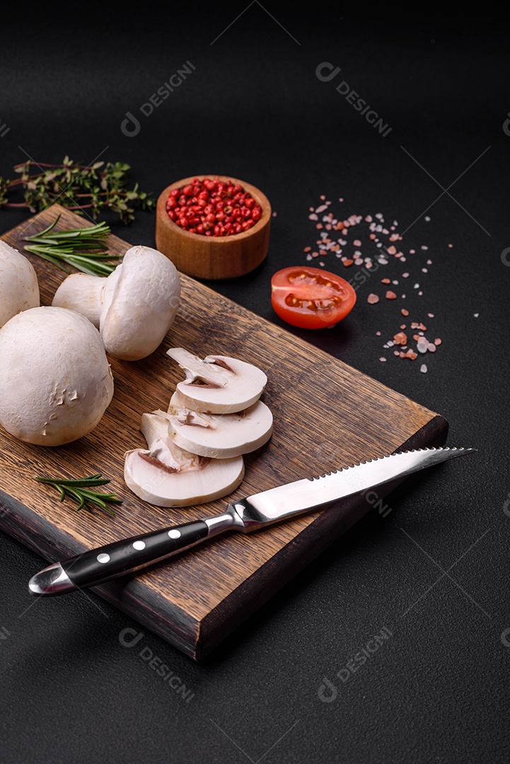 Cogumelos champignon crus frescos sobre uma tábua de madeira com especiarias e ervas sobre um fundo escuro de concreto