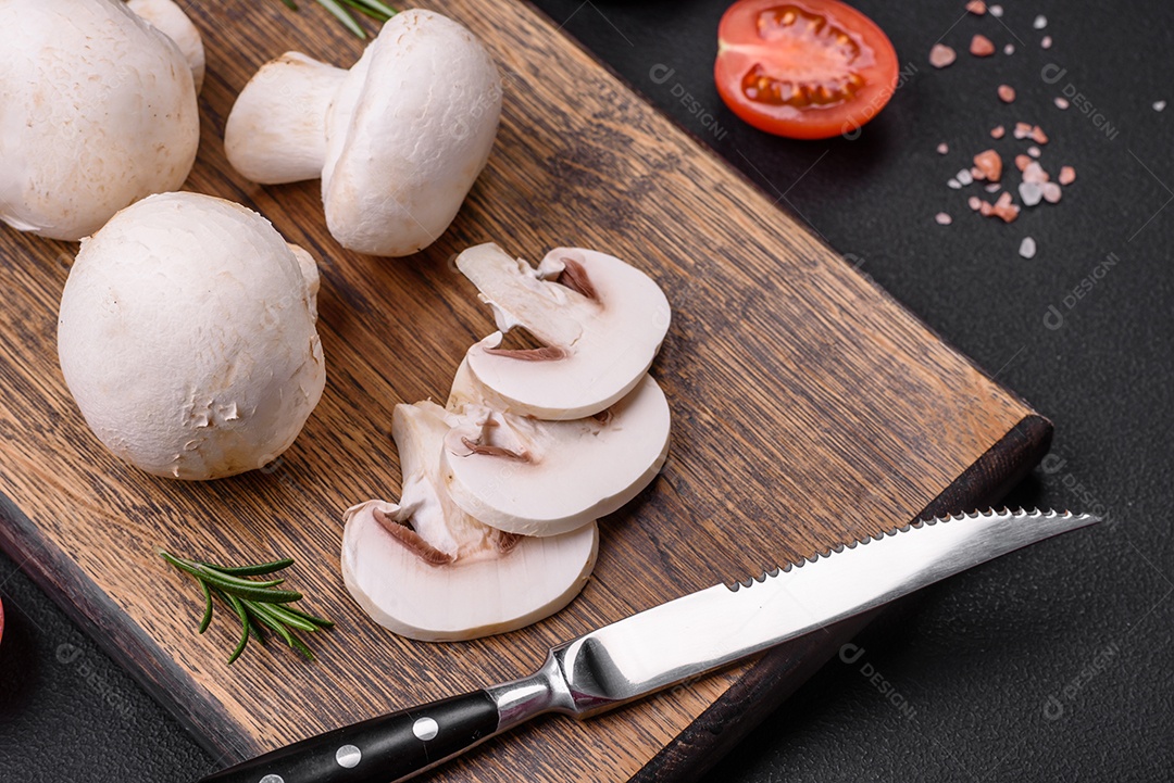 Cogumelos champignon crus frescos sobre uma tábua de madeira com especiarias e ervas sobre um fundo escuro de concreto