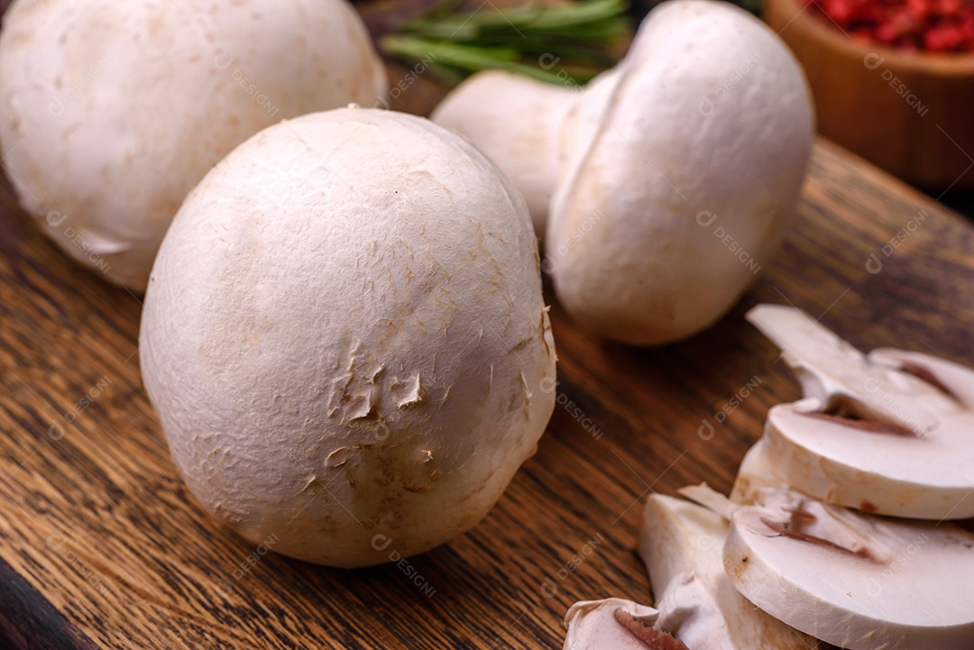 Cogumelos champignon crus frescos sobre uma tábua de madeira com especiarias e ervas sobre um fundo escuro de concreto