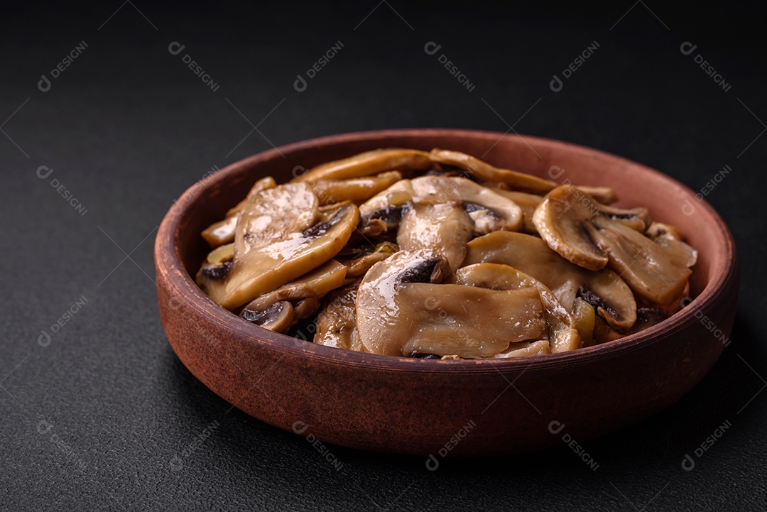 Cogumelos champignon fritos ou cozidos em forma de fatias com cebola num prato de cerâmica marrom sobre um fundo escuro de concreto