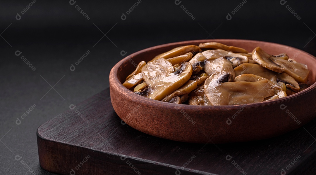 Cogumelos champignon fritos ou cozidos em forma de fatias com cebola num prato de cerâmica marrom sobre um fundo escuro de concreto