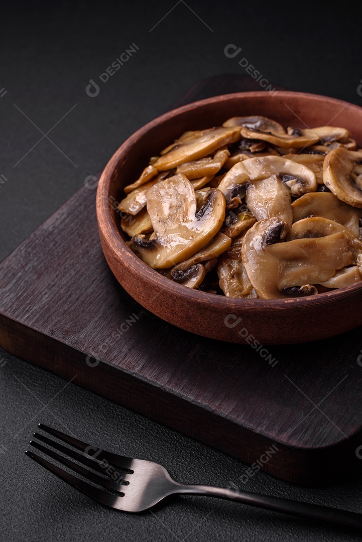 Cogumelos champignon fritos ou cozidos em forma de fatias com cebola num prato de cerâmica marrom sobre um fundo escuro de concreto