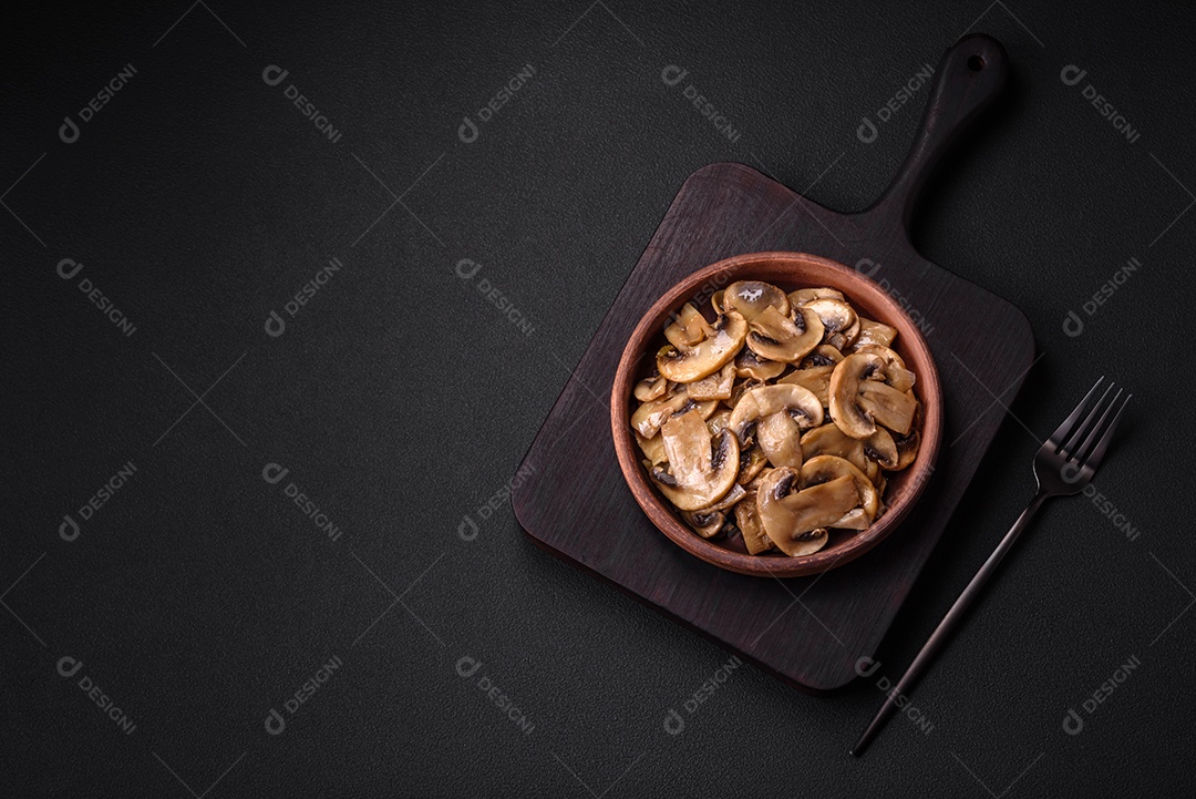 Cogumelos champignon fritos ou cozidos em forma de fatias com cebola num prato de cerâmica marrom sobre um fundo escuro de concreto