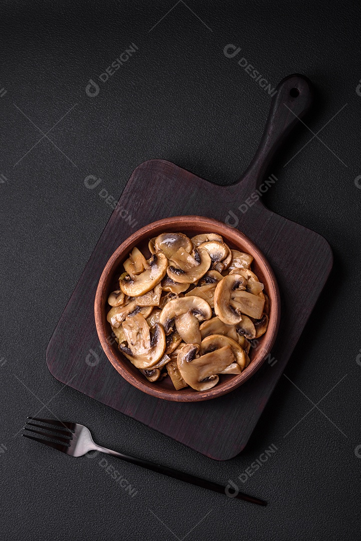 Cogumelos champignon fritos ou cozidos em forma de fatias com cebola num prato de cerâmica marrom sobre um fundo escuro de concreto