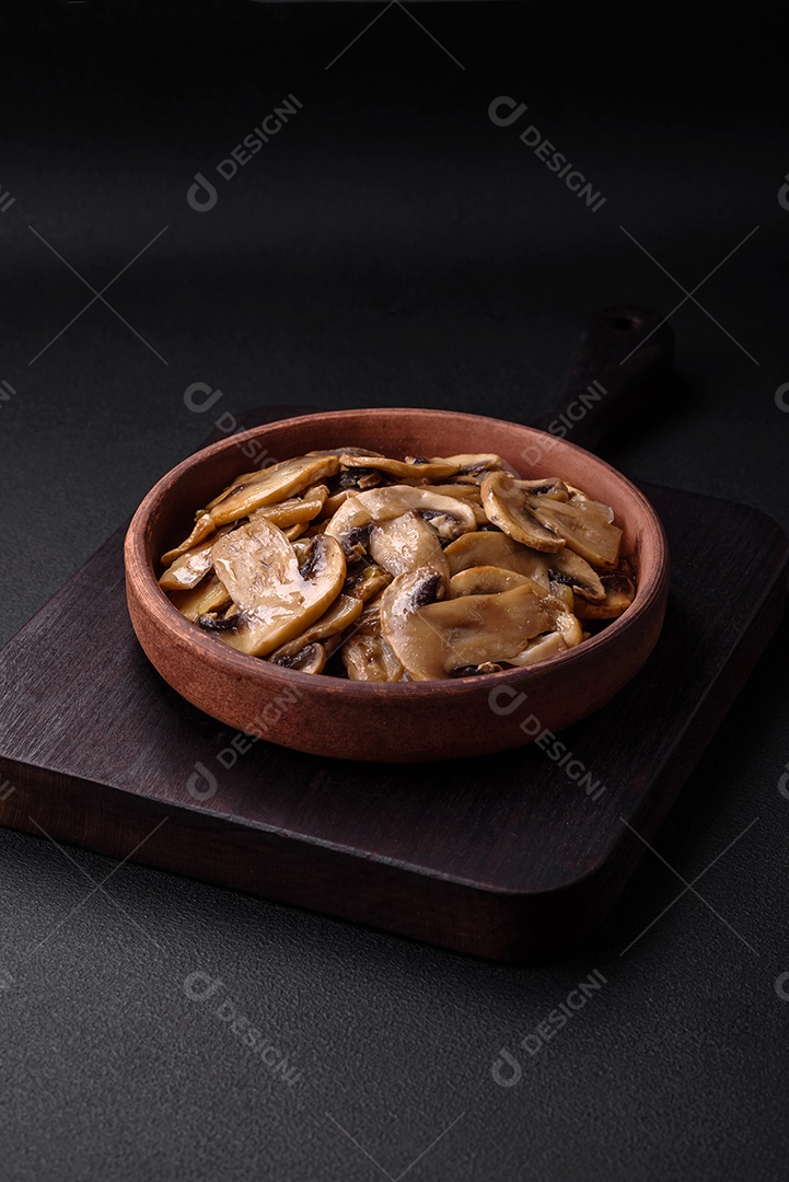 Cogumelos champignon fritos ou cozidos em forma de fatias com cebola num prato de cerâmica marrom sobre um fundo escuro de concreto