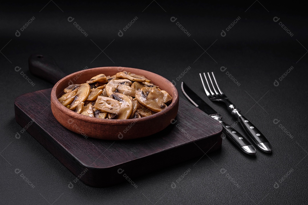 Cogumelos champignon fritos ou cozidos em forma de fatias com cebola num prato de cerâmica marrom sobre um fundo escuro de concreto