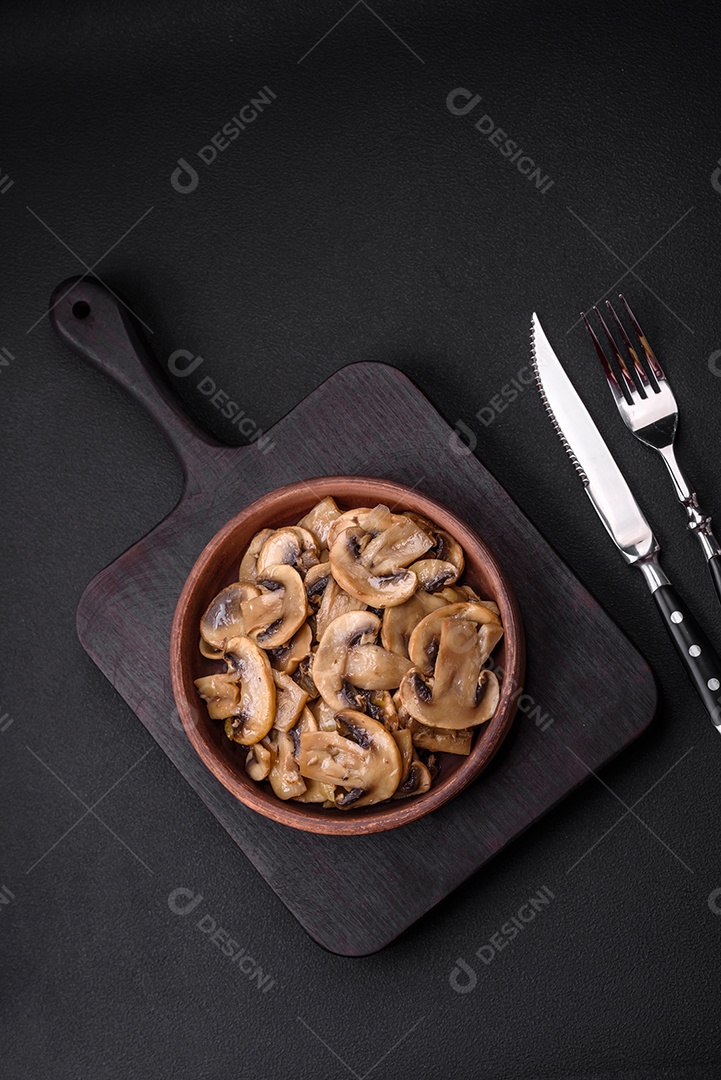 Cogumelos champignon fritos ou cozidos em forma de fatias com cebola num prato de cerâmica marrom sobre um fundo escuro de concreto