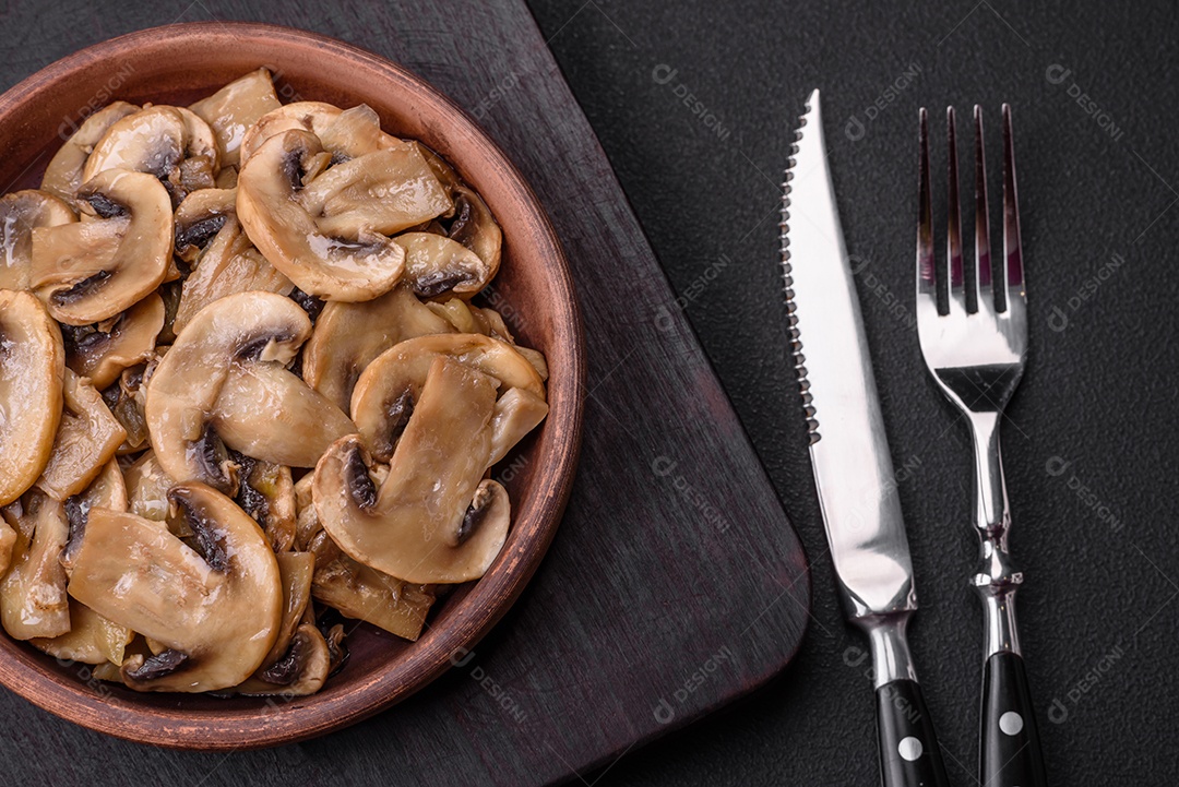 Cogumelos champignon fritos ou cozidos em forma de fatias com cebola num prato de cerâmica marrom sobre um fundo escuro de concreto