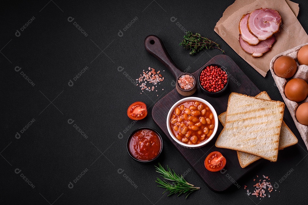 Delicioso café da manhã inglês nutritivo com ovos fritos, tomates, bacon e feijão em um fundo escuro de concreto