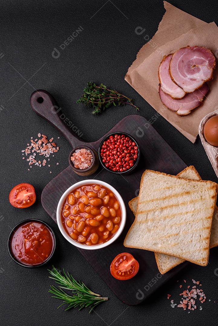 Delicioso café da manhã inglês nutritivo com ovos fritos, tomates, bacon e feijão em um fundo escuro de concreto