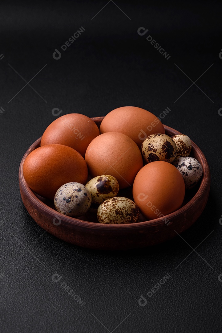 Ovos de galinha e codorna em uma tigela de cerâmica marrom sobre um fundo escuro de concreto.