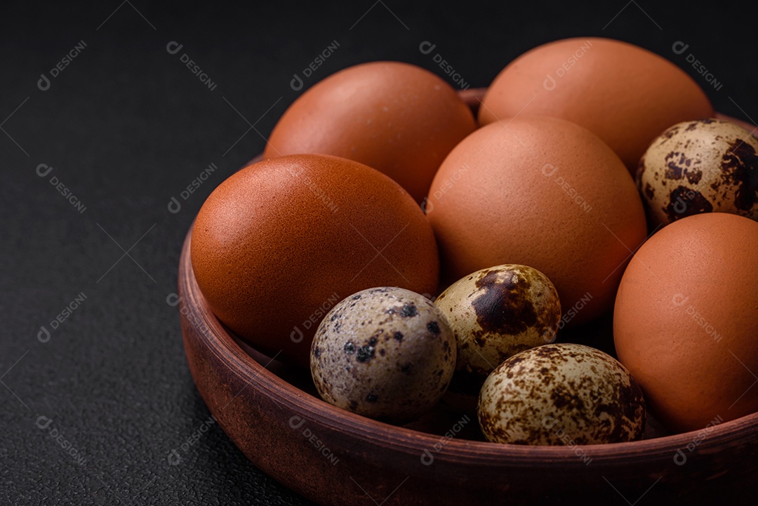 Ovos de galinha e codorna em uma tigela de cerâmica marrom sobre um fundo escuro de concreto.