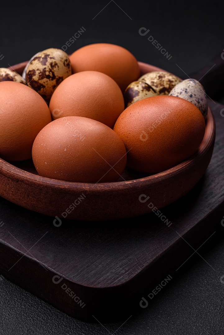 Ovos de galinha e codorna em uma tigela de cerâmica marrom sobre um fundo escuro de concreto.