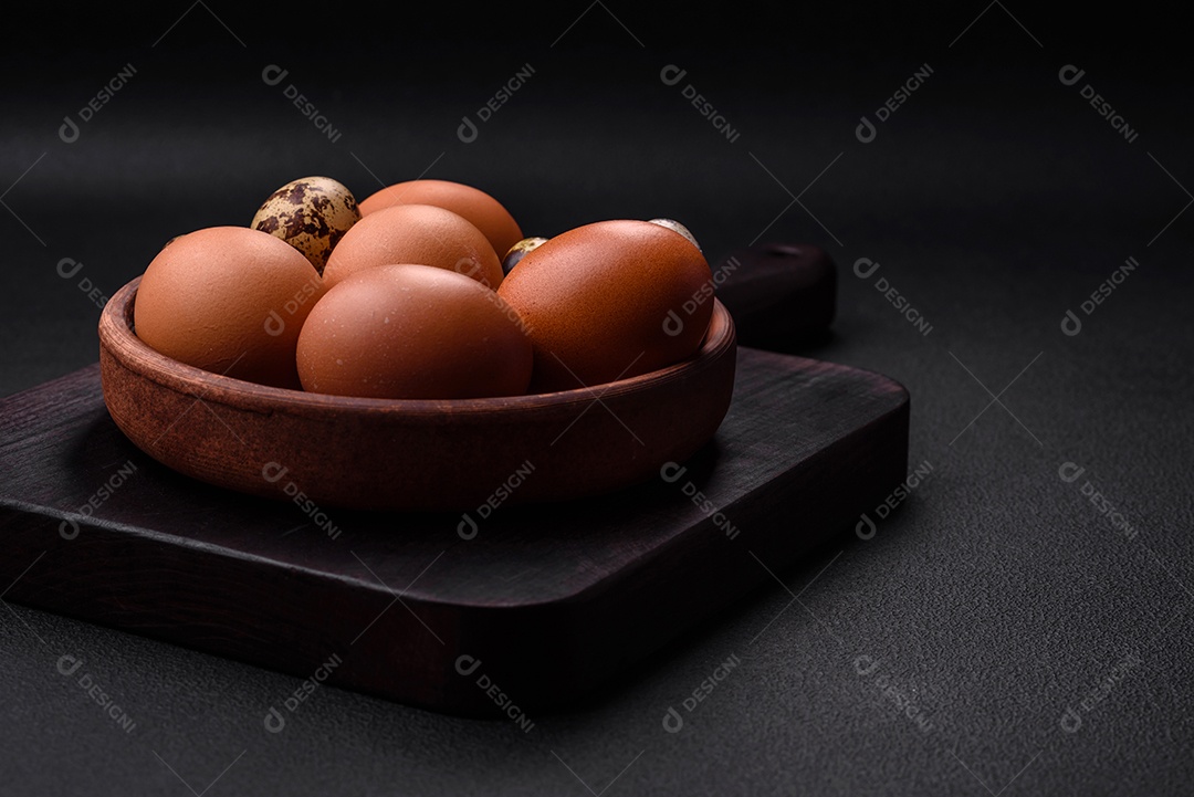 Ovos de galinha e codorna em uma tigela de cerâmica marrom sobre um fundo escuro de concreto.