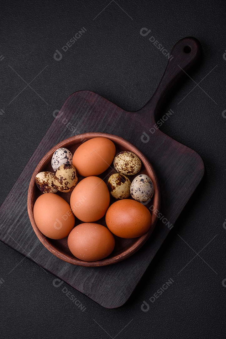 Ovos de galinha e codorna em uma tigela de cerâmica marrom sobre um fundo escuro de concreto.