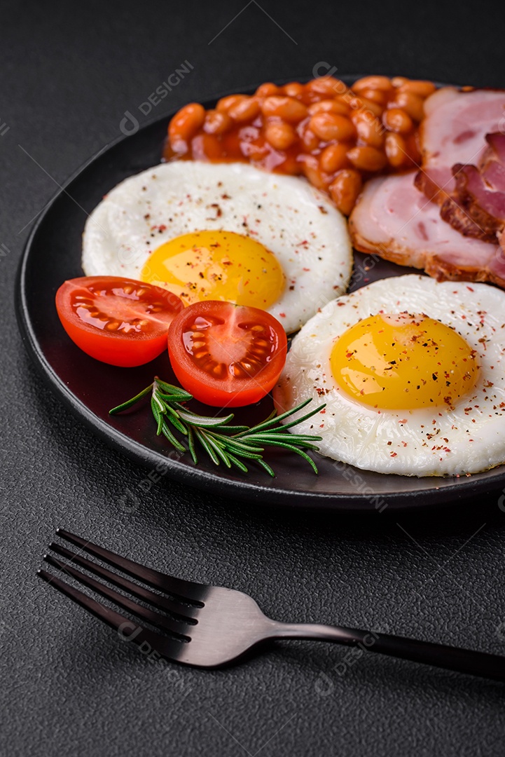 Delicioso café da manhã inglês nutritivo com ovos fritos, tomates, bacon e feijão em um fundo escuro de concreto