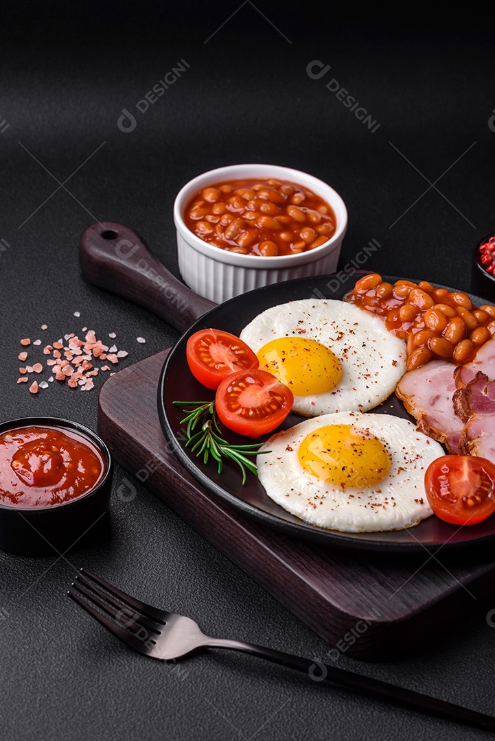 Delicioso café da manhã inglês nutritivo com ovos fritos, tomates, bacon e feijão em um fundo escuro de concreto