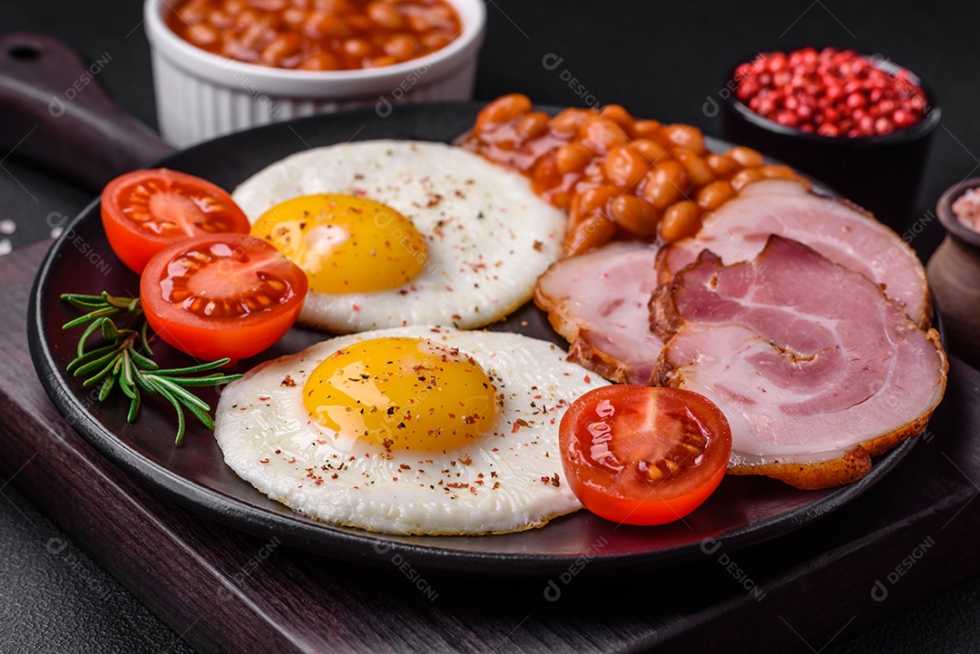 Delicioso café da manhã inglês nutritivo com ovos fritos, tomates, bacon e feijão em um fundo escuro de concreto