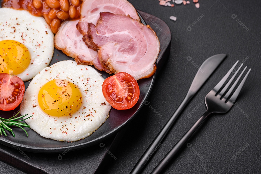 Delicioso café da manhã inglês nutritivo com ovos fritos, tomates, bacon e feijão em um fundo escuro de concreto