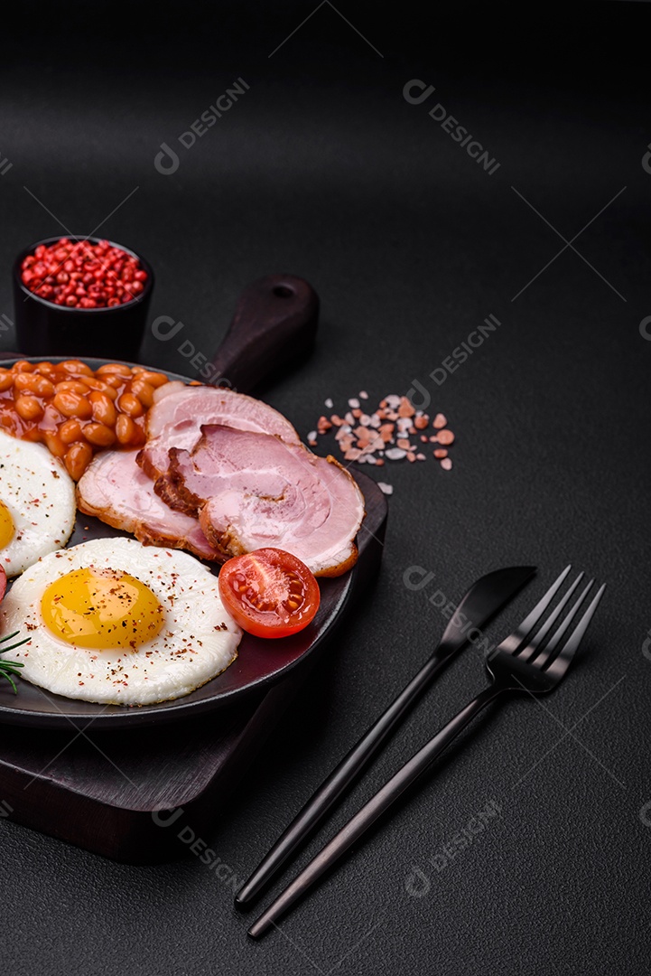 Delicioso café da manhã inglês nutritivo com ovos fritos, tomates, bacon e feijão em um fundo escuro de concreto