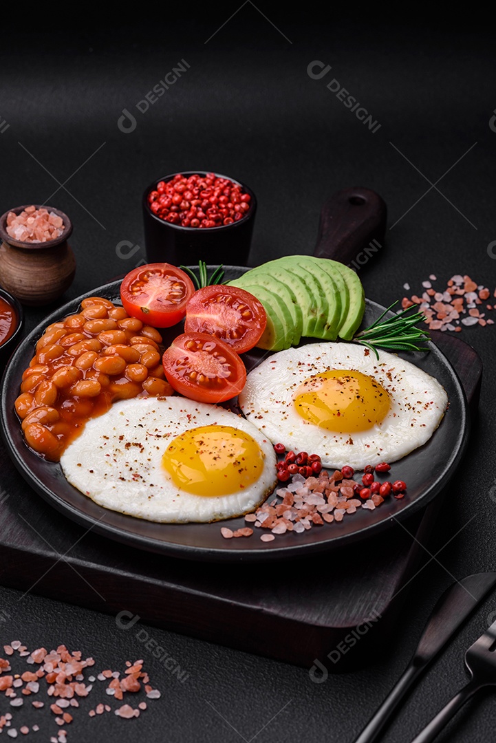 Delicioso café da manhã inglês nutritivo com ovos fritos, tomates, bacon e feijão em um fundo escuro de concreto