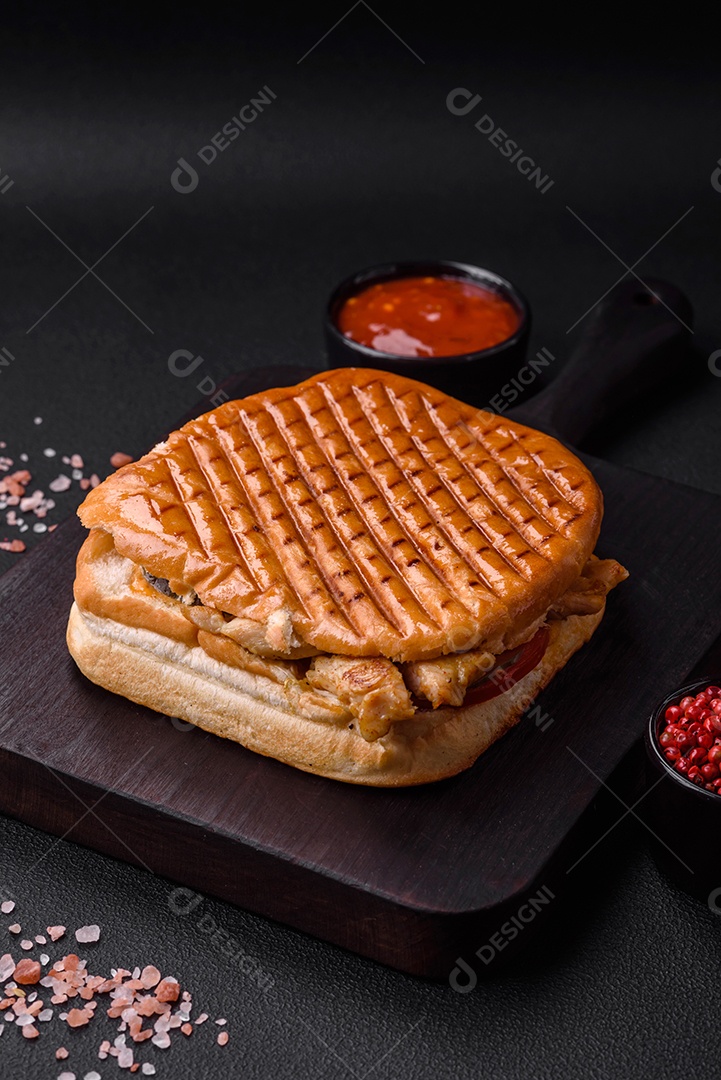 Delicioso sanduíche crocante com peito de frango, tomate, ketchup e especiarias em um fundo escuro de concreto