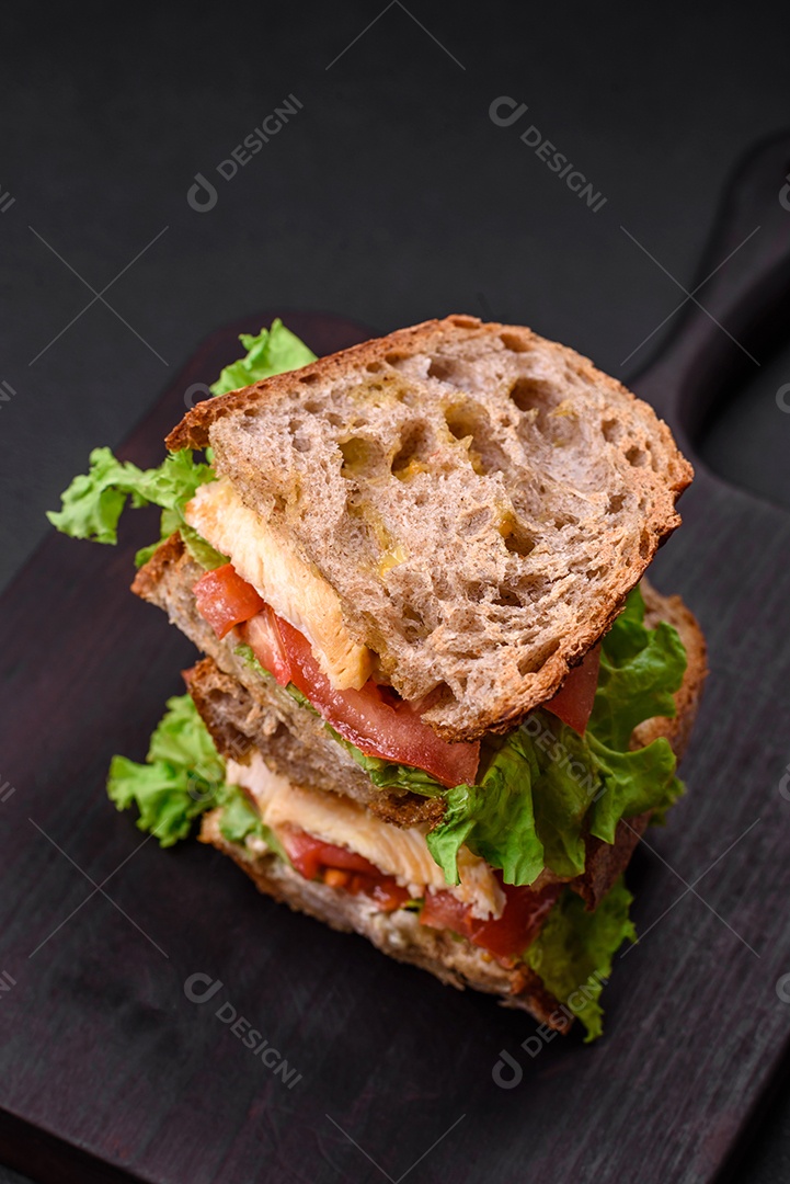 Delicioso sanduíche com torradas crocantes, frango, tomate e alface sobre fundo escuro de concreto