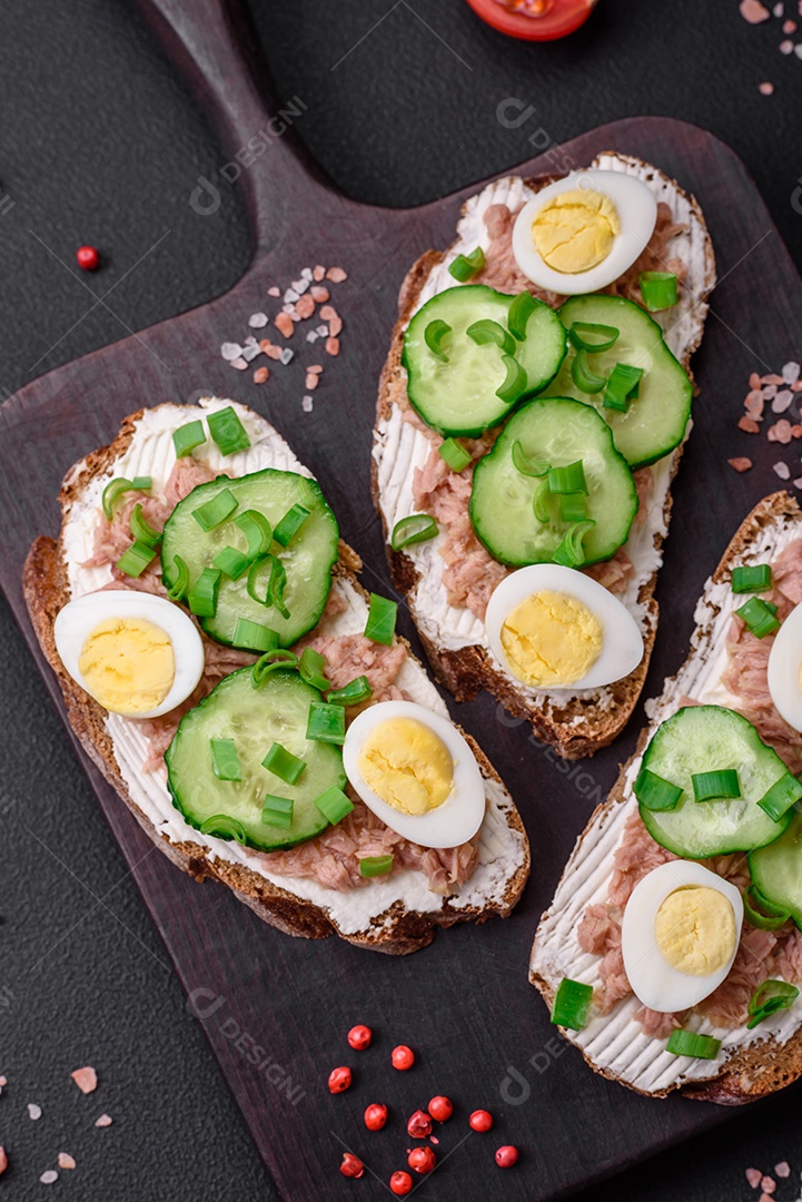 Deliciosos sanduíches compostos por torradas grelhadas, atum enlatado, cream cheese, pepino e ovos de codorna cozidos em um fundo escuro de concreto