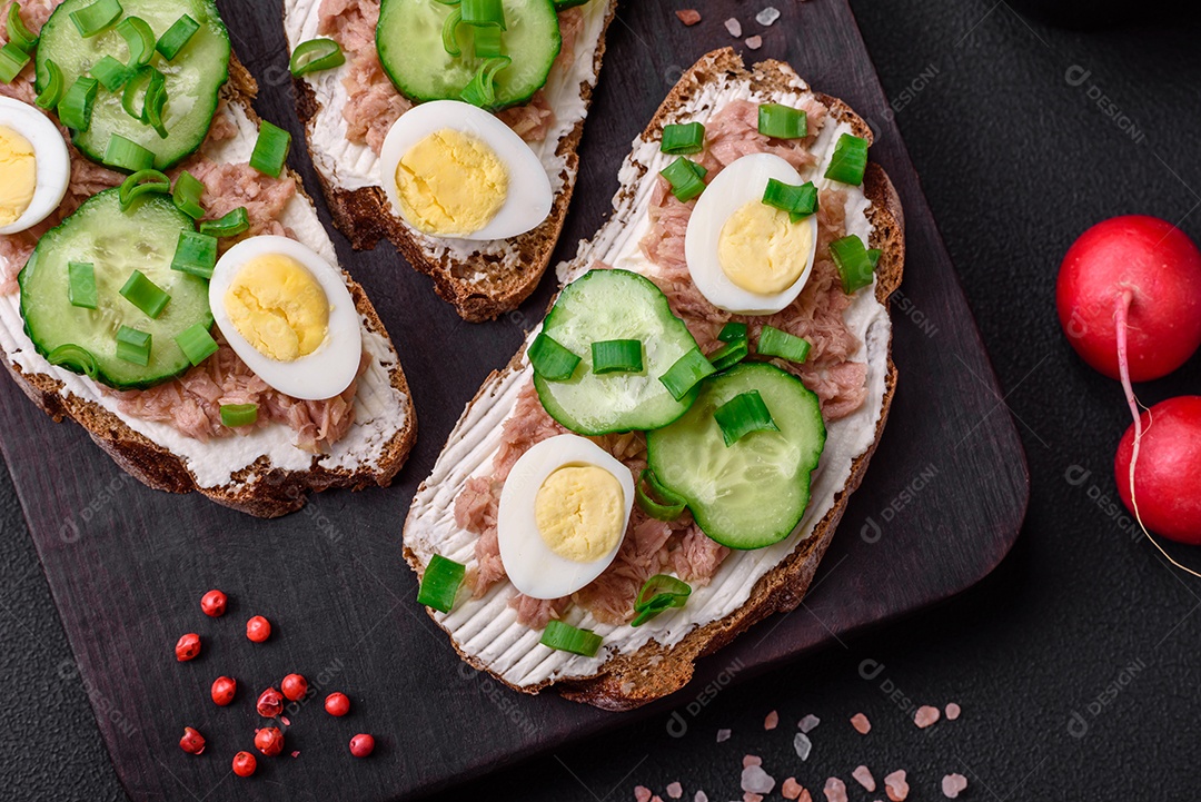 Deliciosos sanduíches compostos por torradas grelhadas, atum enlatado, cream cheese, pepino e ovos de codorna cozidos em um fundo escuro de concreto