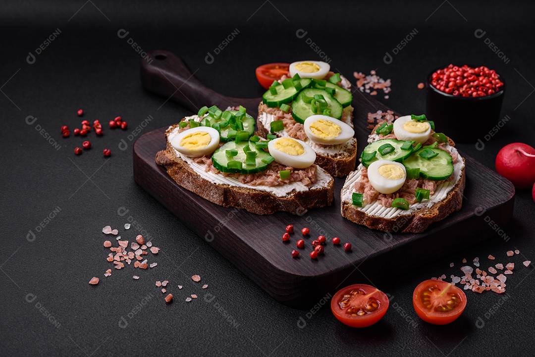 Deliciosos sanduíches compostos por torradas grelhadas, atum enlatado, cream cheese, pepino e ovos de codorna cozidos em um fundo escuro de concreto
