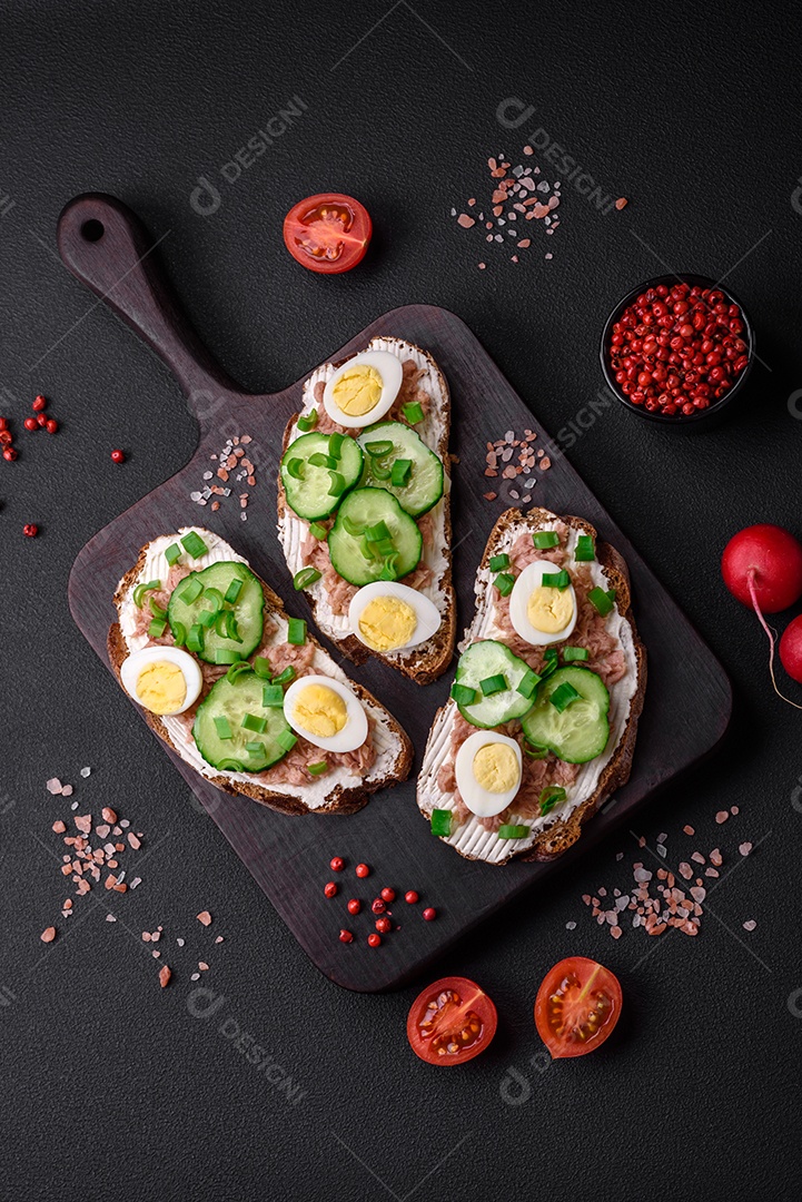 Deliciosos sanduíches compostos por torradas grelhadas, atum enlatado, cream cheese, pepino e ovos de codorna cozidos em um fundo escuro de concreto