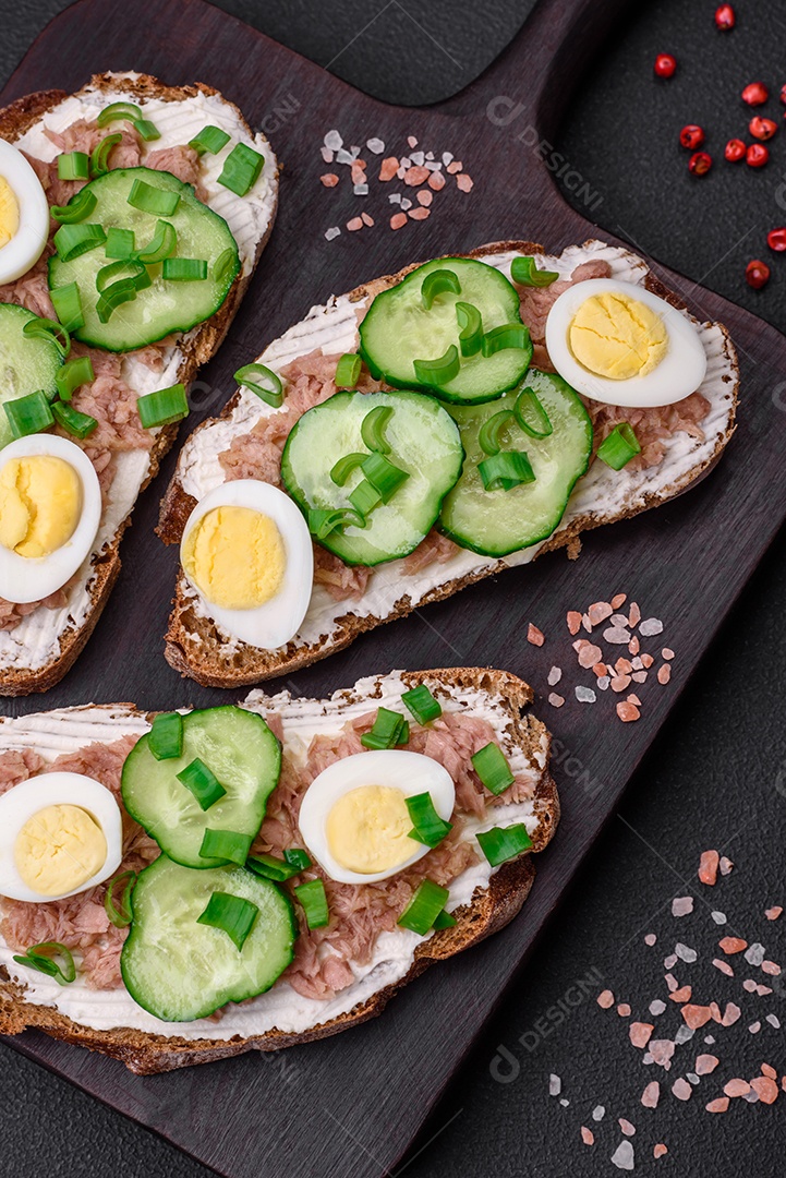 Deliciosos sanduíches compostos por torradas grelhadas, atum enlatado, cream cheese, pepino e ovos de codorna cozidos em um fundo escuro de concreto