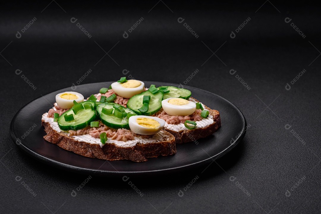 Deliciosos sanduíches compostos por torradas grelhadas, atum enlatado, cream cheese, pepino e ovos de codorna cozidos em um fundo escuro de concreto