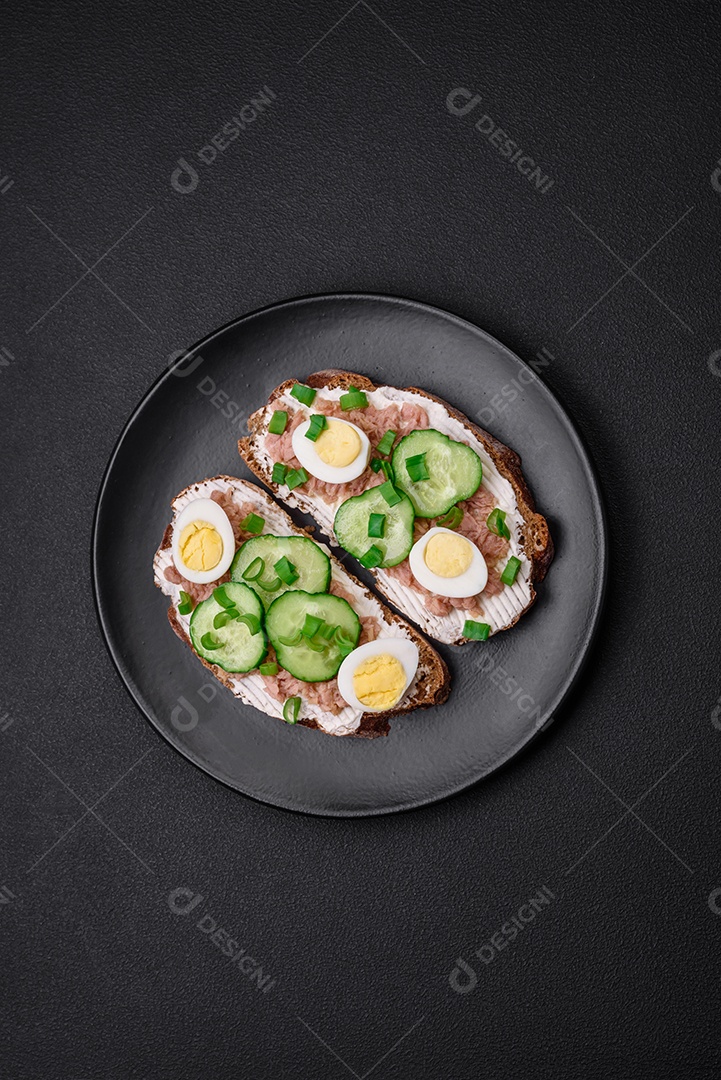 Deliciosos sanduíches compostos por torradas grelhadas, atum enlatado, cream cheese, pepino e ovos de codorna cozidos em um fundo escuro de concreto