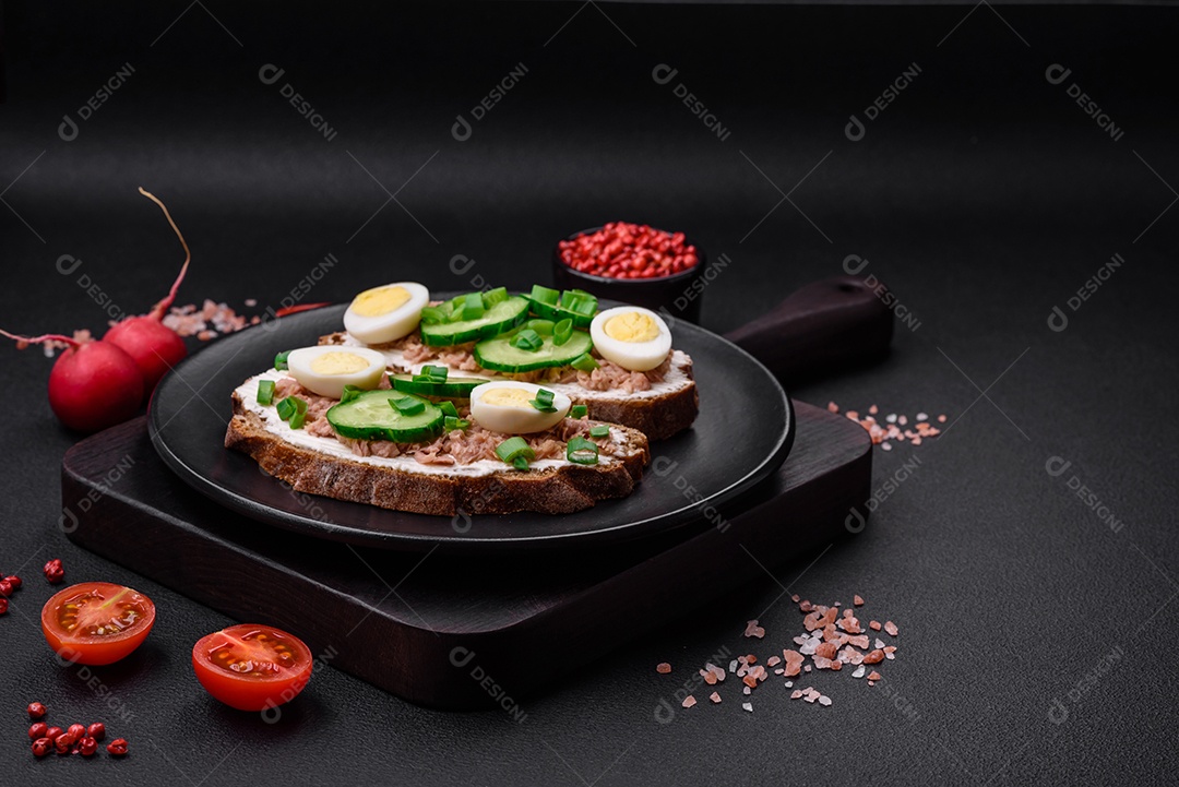 Deliciosos sanduíches compostos por torradas grelhadas, atum enlatado, cream cheese, pepino e ovos de codorna cozidos em um fundo escuro de concreto