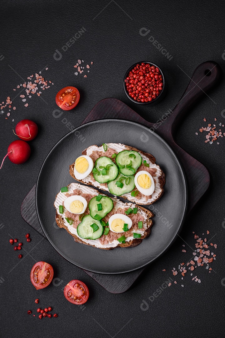 Deliciosos sanduíches compostos por torradas grelhadas, atum enlatado, cream cheese, pepino e ovos de codorna cozidos em um fundo escuro de concreto