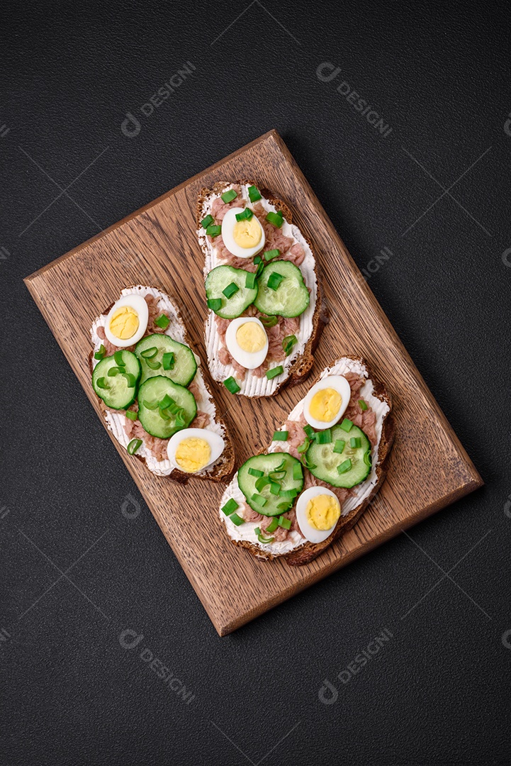 Deliciosos sanduíches compostos por torradas grelhadas, atum enlatado, cream cheese, pepino e ovos de codorna cozidos em um fundo escuro de concreto