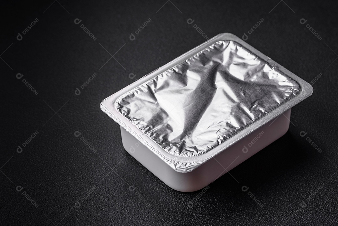 Caixa plástica retangular com iogurte ou queijo hermeticamente selada com tampa de alumínio sobre fundo escuro de concreto