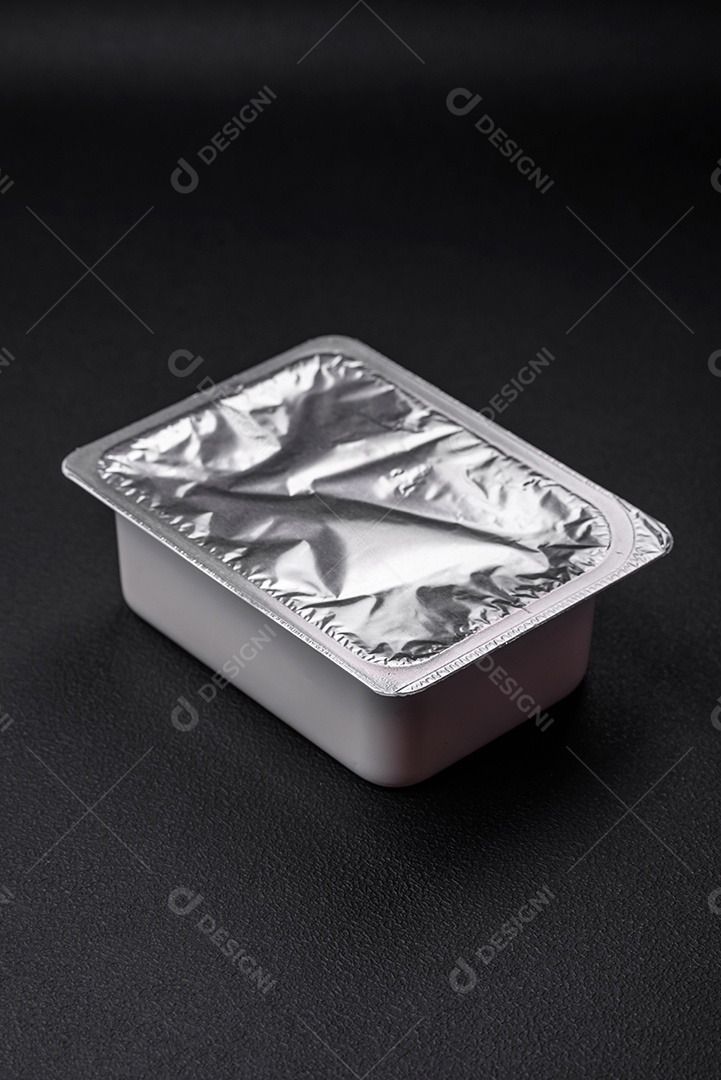 Caixa plástica retangular com iogurte ou queijo hermeticamente selada com tampa de alumínio sobre fundo escuro de concreto