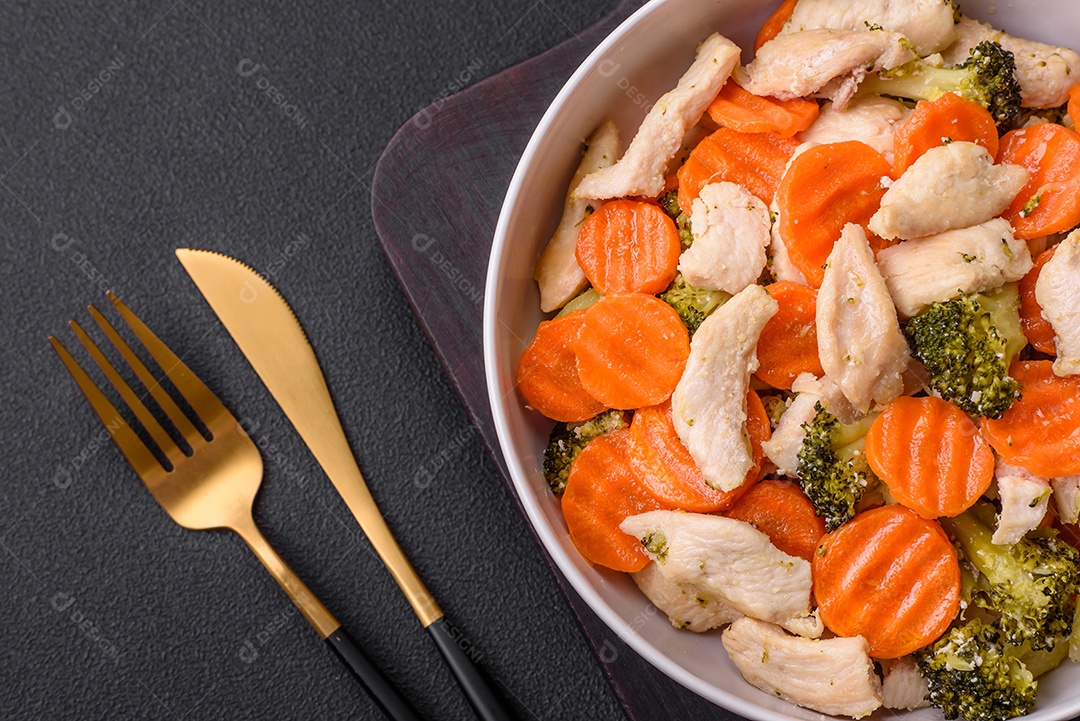 Delicioso prato composto por pedaços de frango cozido, brócolis e cenoura com especiarias e ervas sobre um fundo escuro de concreto