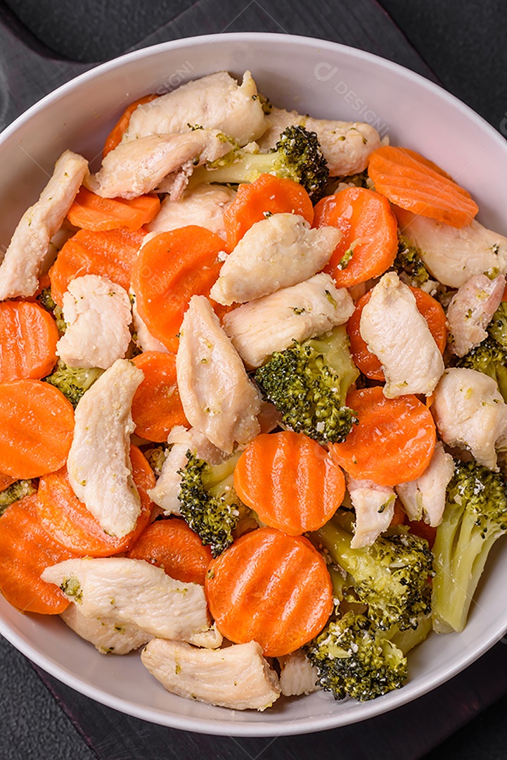 Delicioso prato composto por pedaços de frango cozido, brócolis e cenoura com especiarias e ervas sobre um fundo escuro de concreto