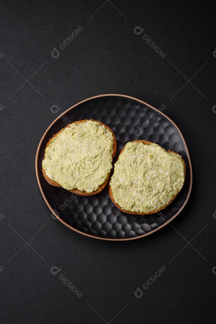 Torradas grelhadas com pasta de abacate, ovos, cream cheese, sal, especiarias e ervas sobre uma mesa de concreto texturizado