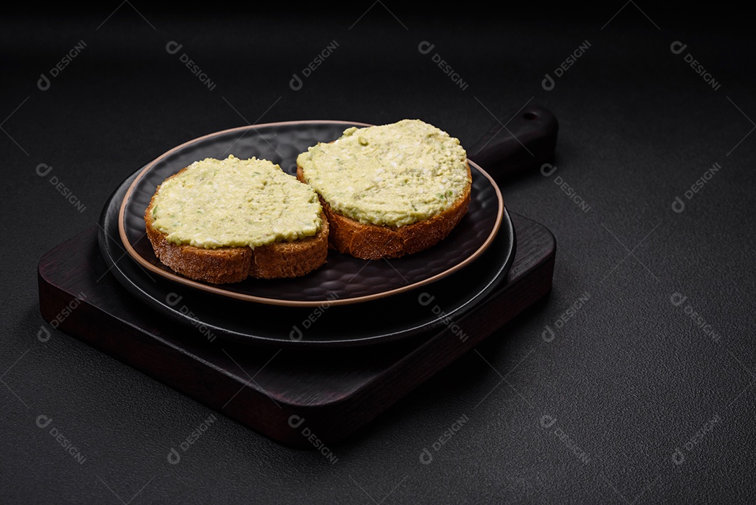 Torradas grelhadas com pasta de abacate, ovos, cream cheese, sal, especiarias e ervas sobre uma mesa de concreto texturizado