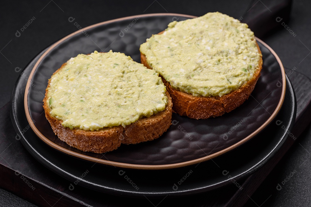 Torradas grelhadas com pasta de abacate, ovos, cream cheese, sal, especiarias e ervas sobre uma mesa de concreto texturizado