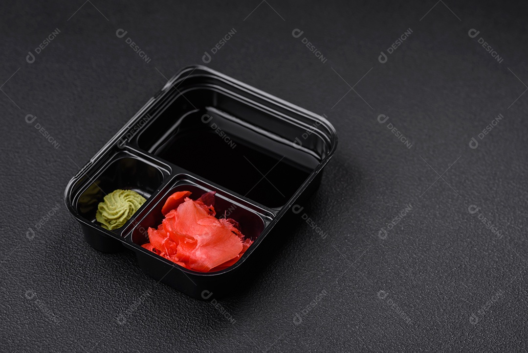 Gengibre em conserva, molho de soja e wasabi em recipientes de plástico porcionados sobre um fundo escuro de concreto