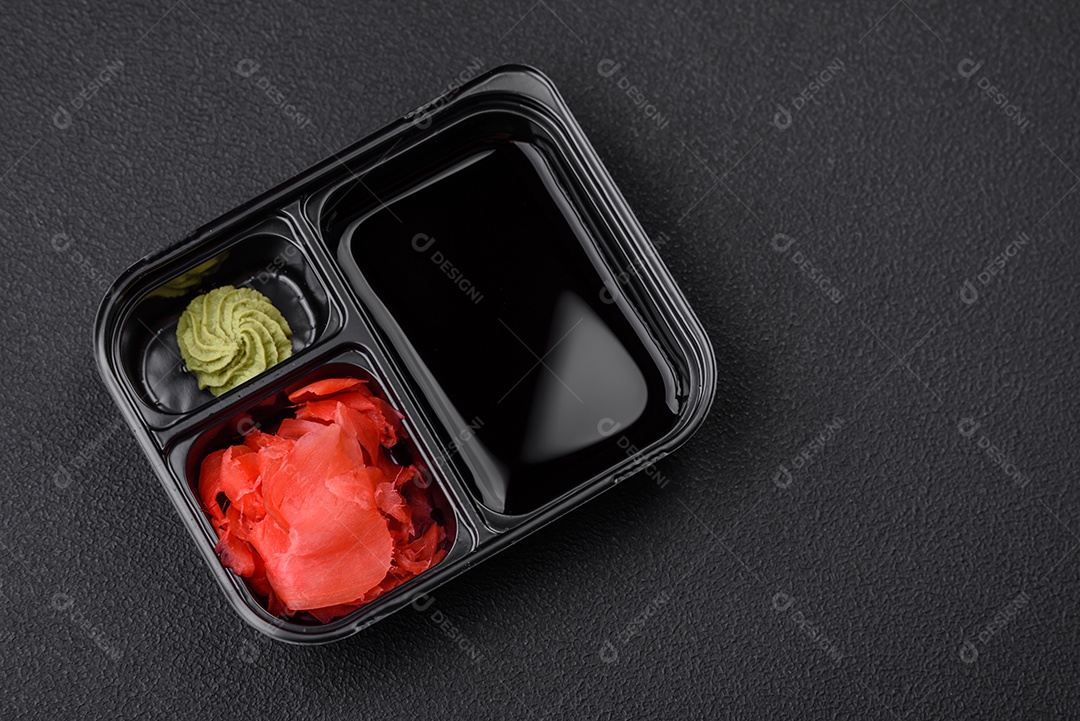 Gengibre em conserva, molho de soja e wasabi em recipientes de plástico porcionados sobre um fundo escuro de concreto