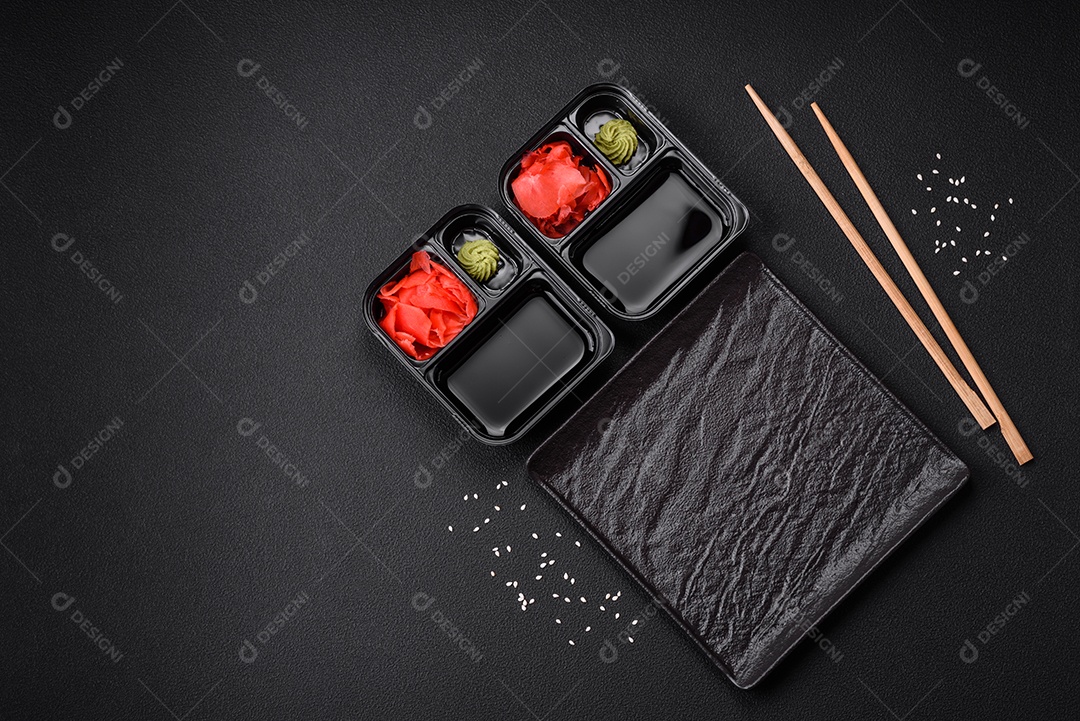 Gengibre em conserva, molho de soja e wasabi em recipientes de plástico porcionados sobre um fundo escuro de concreto