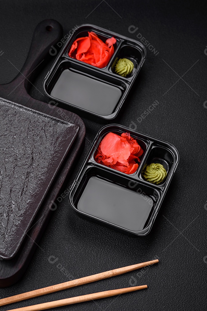 Gengibre em conserva, molho de soja e wasabi em recipientes de plástico porcionados sobre um fundo escuro de concreto